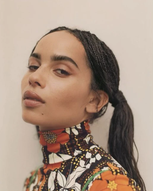 Close-up portrait of a woman with dark braided hair, neutral makeup, and wearing a high-neck floral top, against a light backdrop.