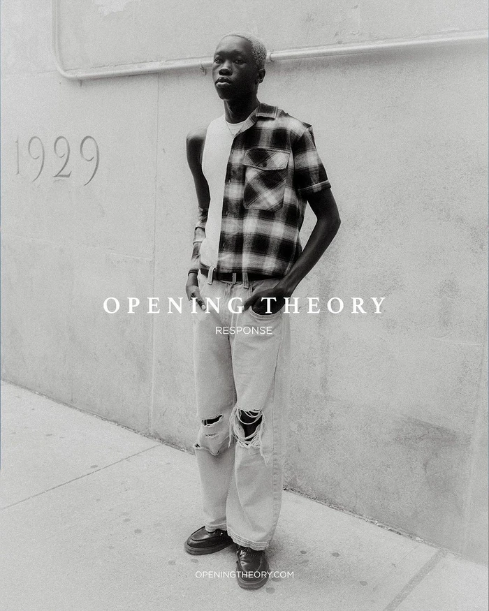 Black man wearing a plaid shirt and distressed jeans standing against a textured concrete wall with overlaid text