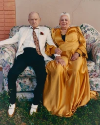 Elderly couple seated together on a floral sofa outdoors, wearing formal attire including a white suit and a golden gown.