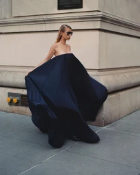 Model in a voluminous dark strapless pleated gown and sunglasses walking past a stone building exterior by Alexander Saladrigas