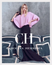 Model in pink blouse and dark maxi skirt standing on a teal velvet sofa against a grey textured wall.