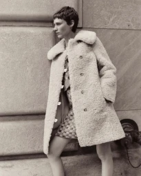 Model with short hair wearing a fuzzy textured coat leaning against a stone building wall in New York.
