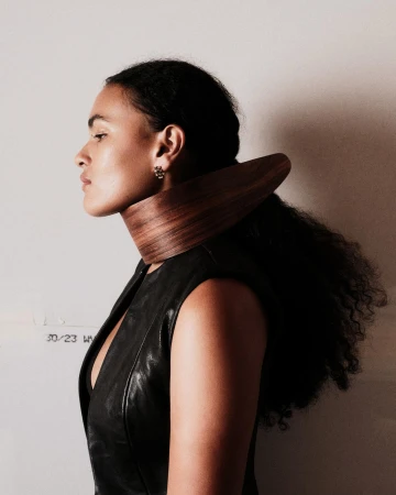 Profile portrait of a woman wearing a dark sleeveless top and a curved wooden neckpiece against a plain wall.