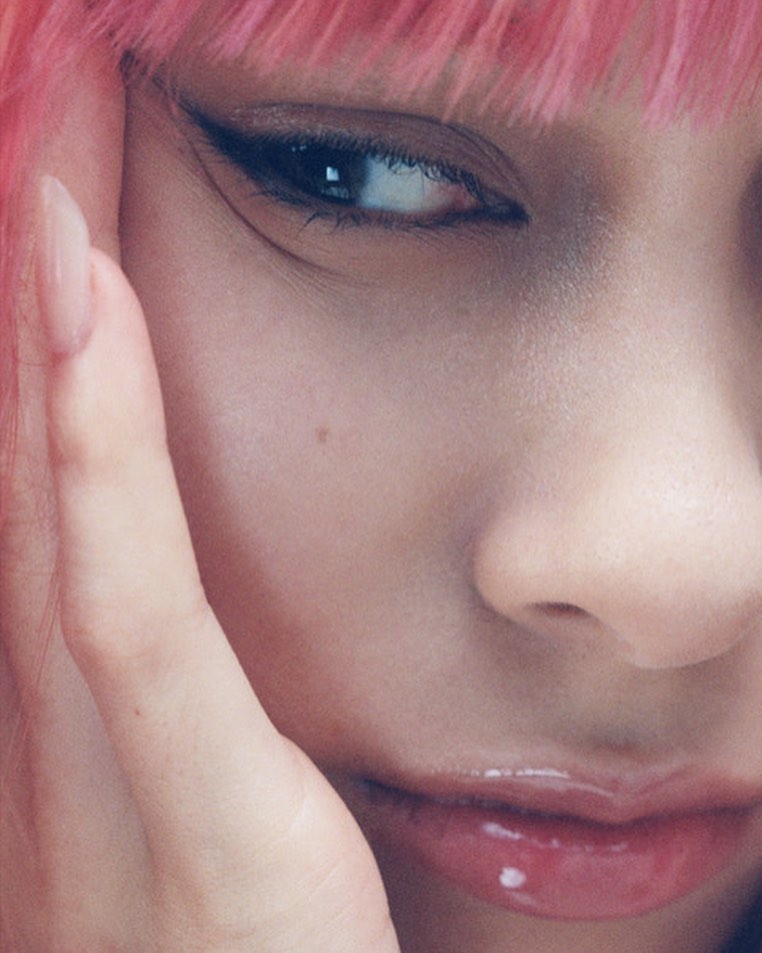Close-up of a woman with pink hair and winged eyeliner, showcasing beauty and makeup details against a blurred background.