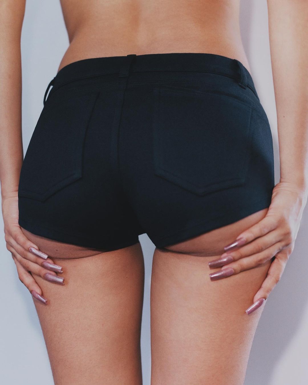 Close-up studio shot of an adult female's lower back, hips, and thighs wearing black jean shorts. She touches the back of her thighs.