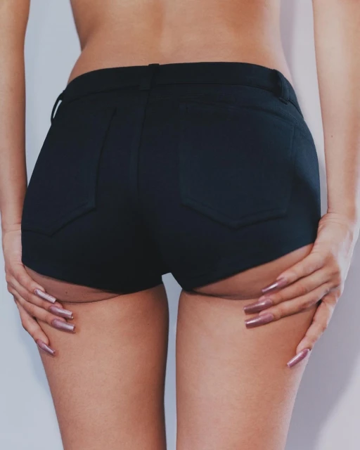Close-up studio shot of an adult female's lower back, hips, and thighs wearing black jean shorts. She touches the back of her thighs.
