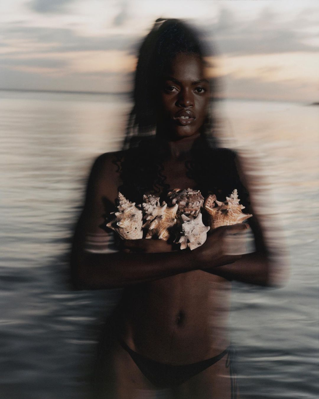 An African American woman stands in the water with seashells covering her chest at dusk, exuding a serene and natural beauty.