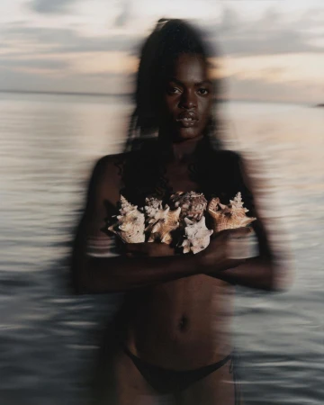 An African American woman stands in the water with seashells covering her chest at dusk, exuding a serene and natural beauty.
