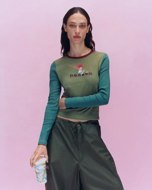 A fair-skinned young woman poses in a fashion shot. She is wearing a green long-sleeved t-shirt, green cargo pants, and holding a cup.