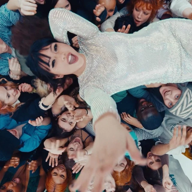 Eye-level view of a woman with dark hair lying among a large group of diverse people, all facing the camera in an indoor setting.