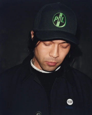 Portrait of a man wearing a black cap with a green PIL logo and a dark jacket, photographed by Alexandra Leese.