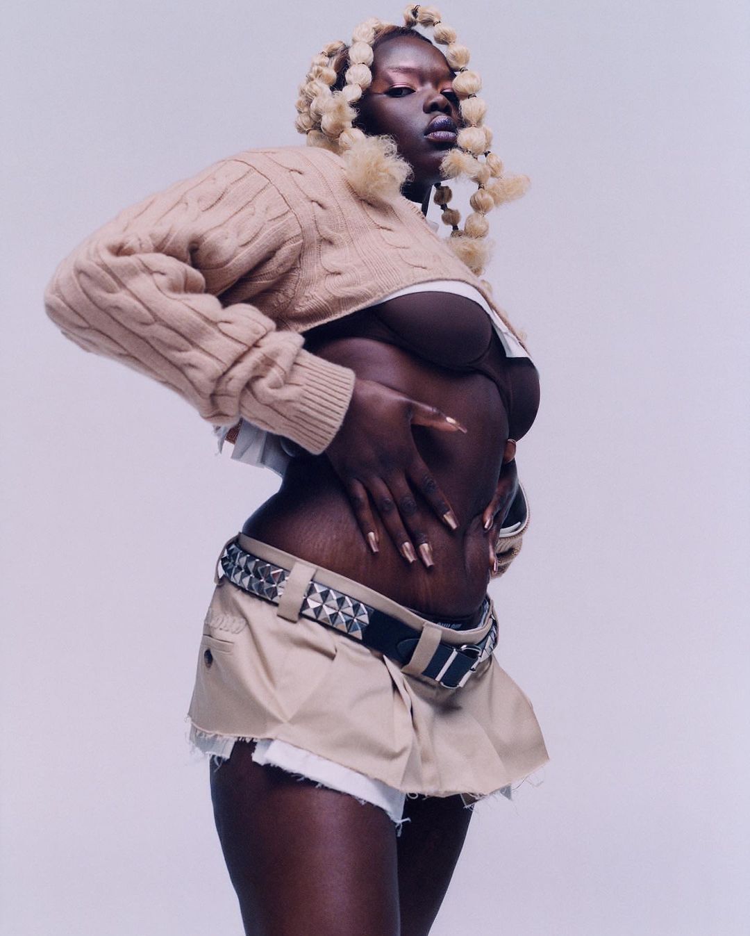 Fashion portrait of an adult woman with dark skin and blonde braided hair wearing a cropped sweater, bra, and short skirt in a studio.