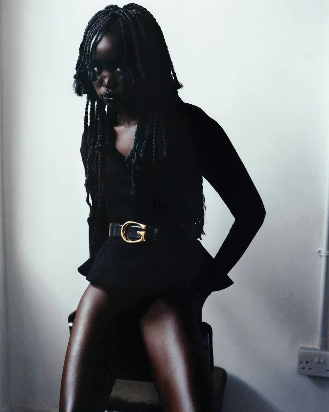 A dark-skinned woman with braided hair, wearing a black top, skirt, and belt, sitting on a chair in front of a white wall with a neutral expression.