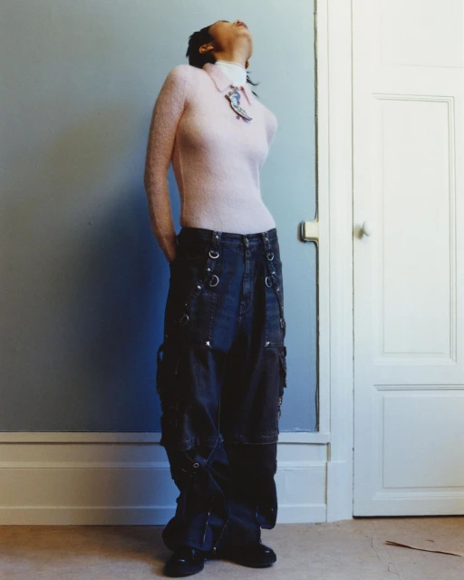 Woman in pink top and dark baggy jeans stands near a white door, looking upward in a studio setting with a painted wall.