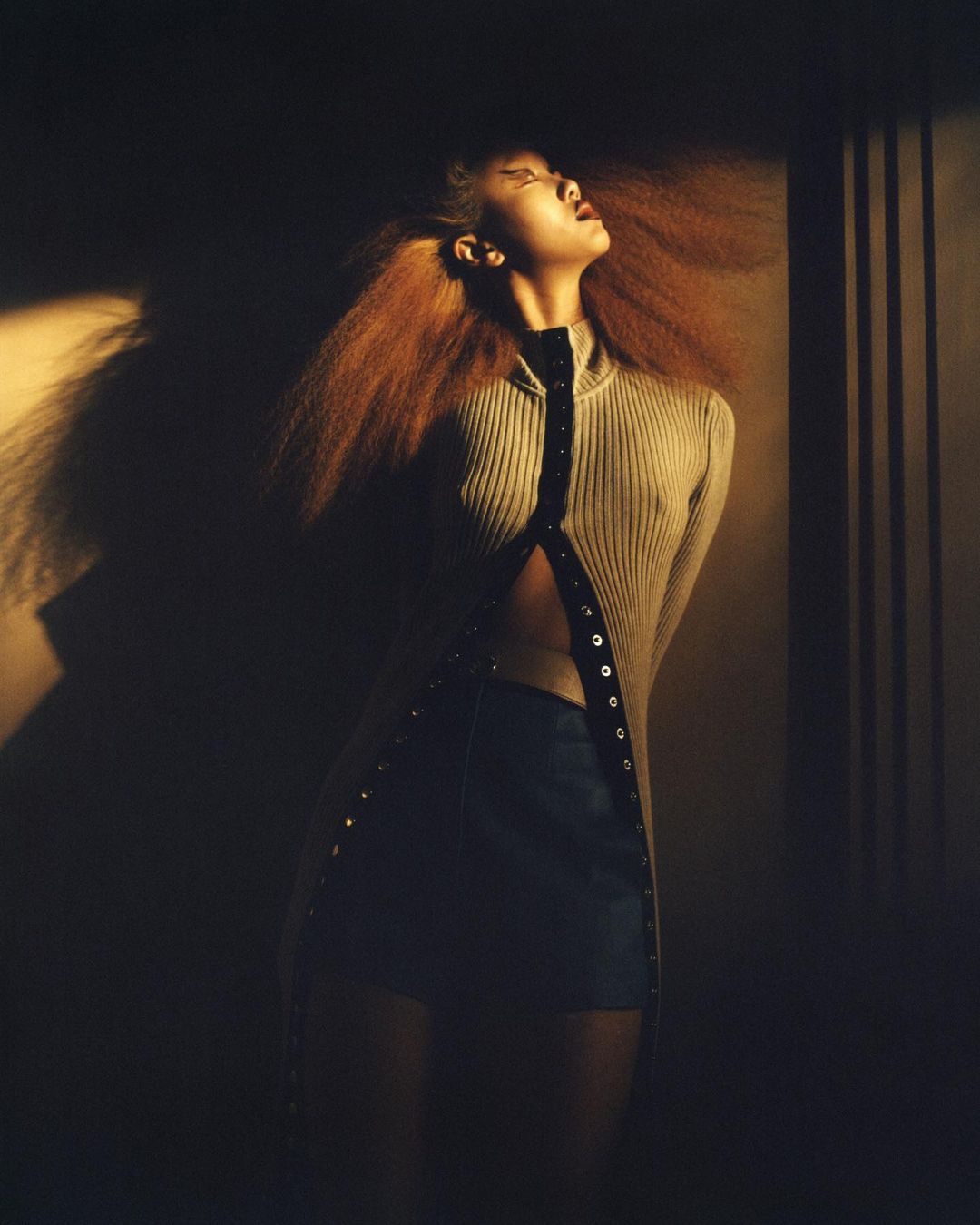 Artistic studio portrait of a red-haired woman in a sweater dress with an open front and dark mini skirt, warm lighting, and shadow.