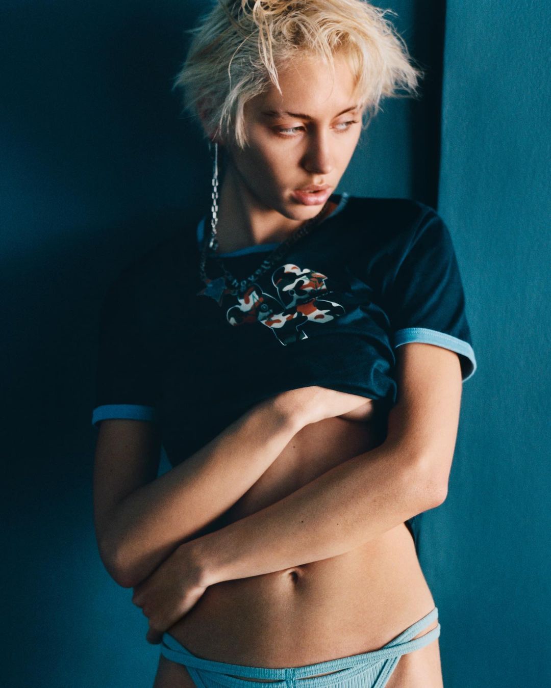 A woman with short blonde hair in a dark shirt and light blue underwear strikes a pose indoors, with a dramatic lighting.