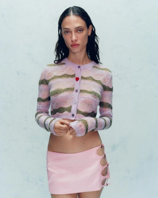 A fashionable woman poses in a studio, wearing a light pink mini skirt and a striped lavender cardigan. Full studio shot. 