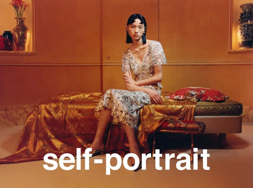 An Asian woman in a sequined dress sits on a bed in a golden-toned room. Vases are displayed in the background, shot for Self-Portrait.