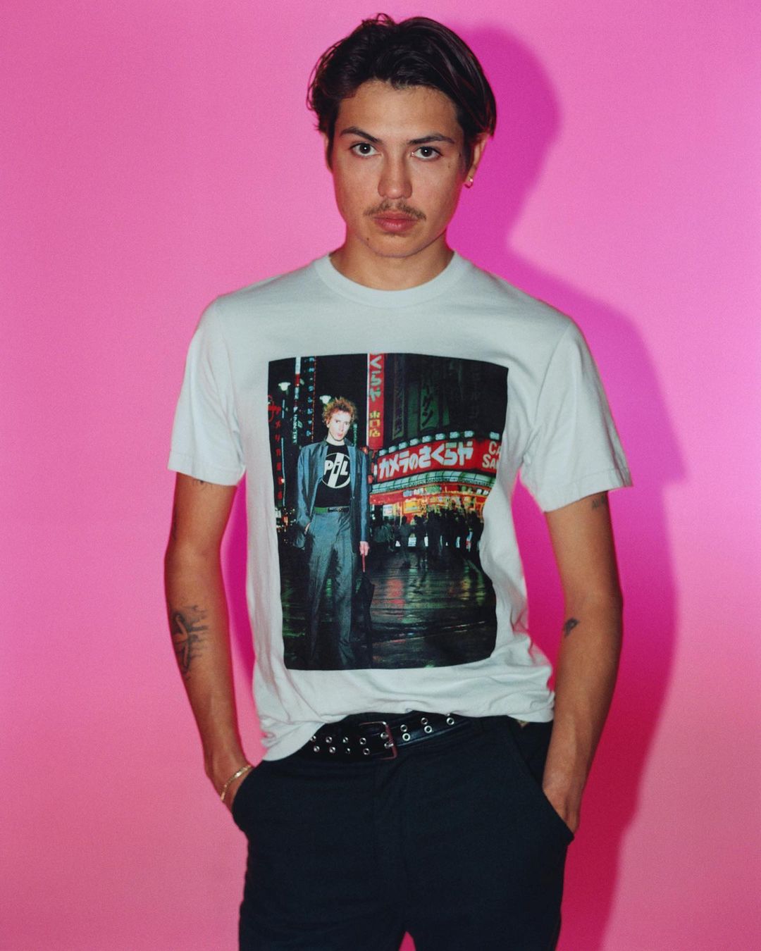 Studio shot of a young adult male wearing a t-shirt with an urban image, black pants and belt, posing against a bright pink background.