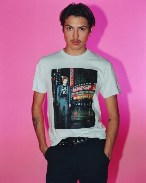 Studio shot of a young adult male wearing a t-shirt with an urban image, black pants and belt, posing against a bright pink background.
