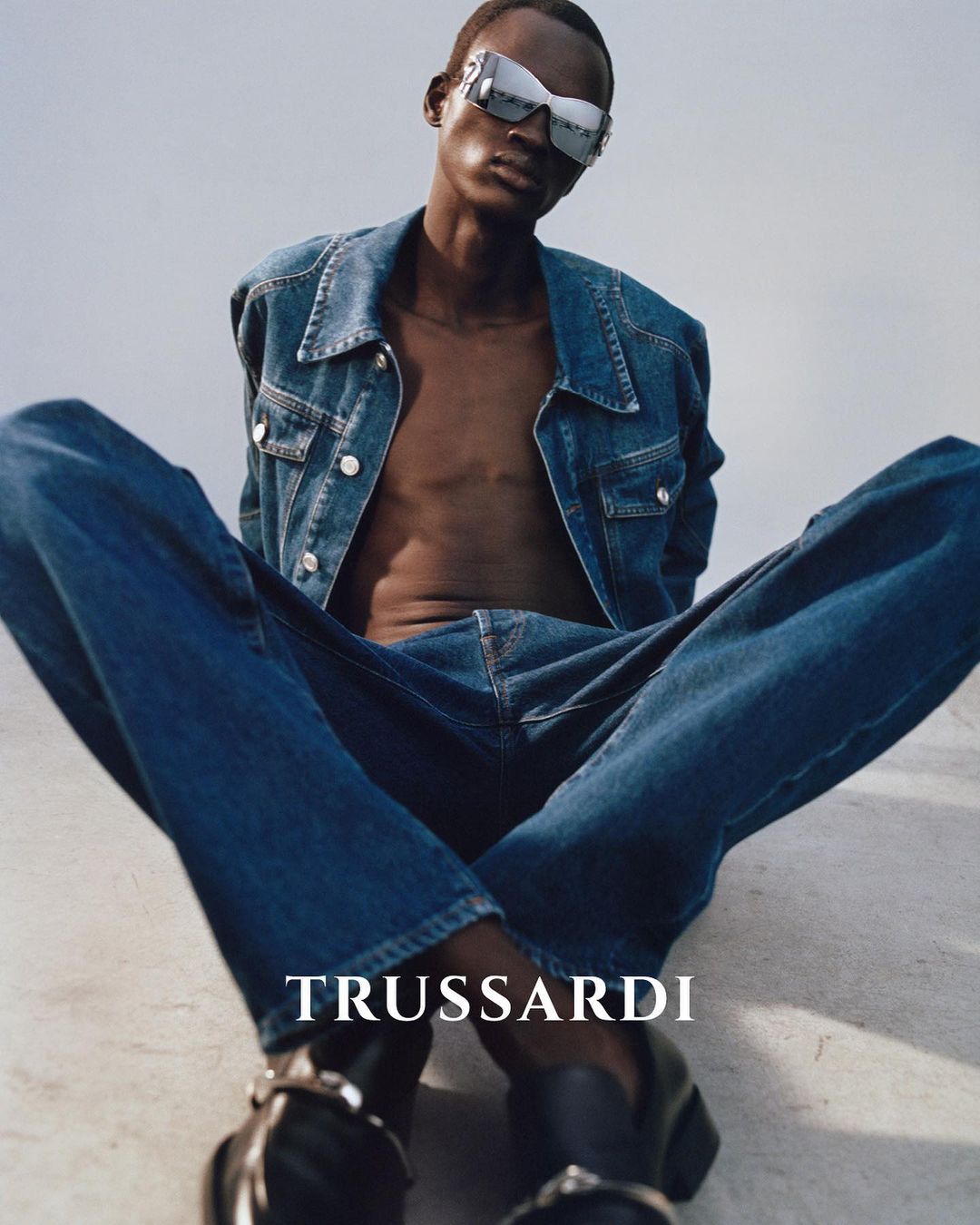 Fashion portrait of an adult male in blue denim jacket and jeans, sunglasses, and black shoes, against a light grey studio backdrop.