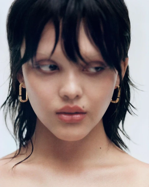 Close-up portrait of a young adult woman with short dark hair, wearing gold earrings, with a neutral facial expression against a bright background.