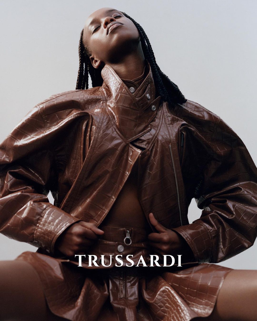 Fashion shot of a Black female model wearing a brown faux-crocodile jacket and mini skirt, posing in a studio with neutral background.