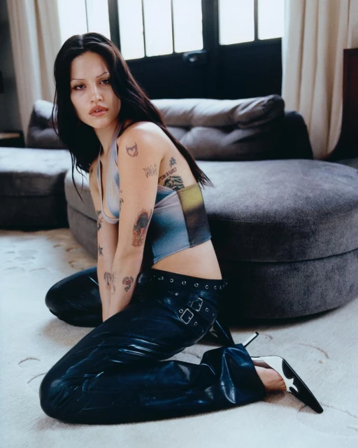 Attractive young woman with dark hair kneels indoors wearing a halter top and shiny black pants, posing against a neutral gray backdrop.