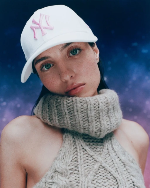 Close-up of a woman with fair skin and green eyes, wearing a white baseball cap with a pink logo and a chunky knit sweater.