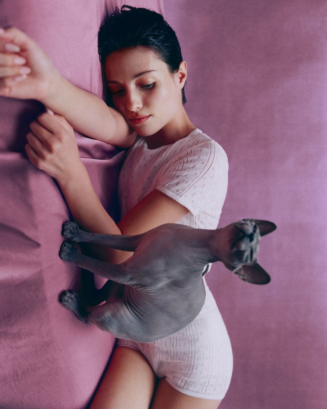 Woman in white loungewear laying on a pink textured surface with a hairless grey cat, horizontal, medium shot