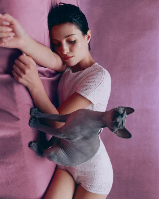 Woman in white loungewear laying on a pink textured surface with a hairless grey cat, horizontal, medium shot