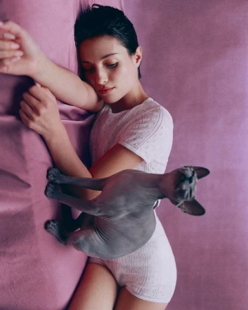 Woman in white loungewear laying on a pink textured surface with a hairless grey cat, horizontal, medium shot