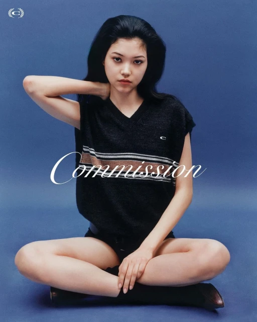 Portrait of an adult female in a sleeveless top, sitting cross-legged against a blue backdrop in a studio, fashion editorial.
