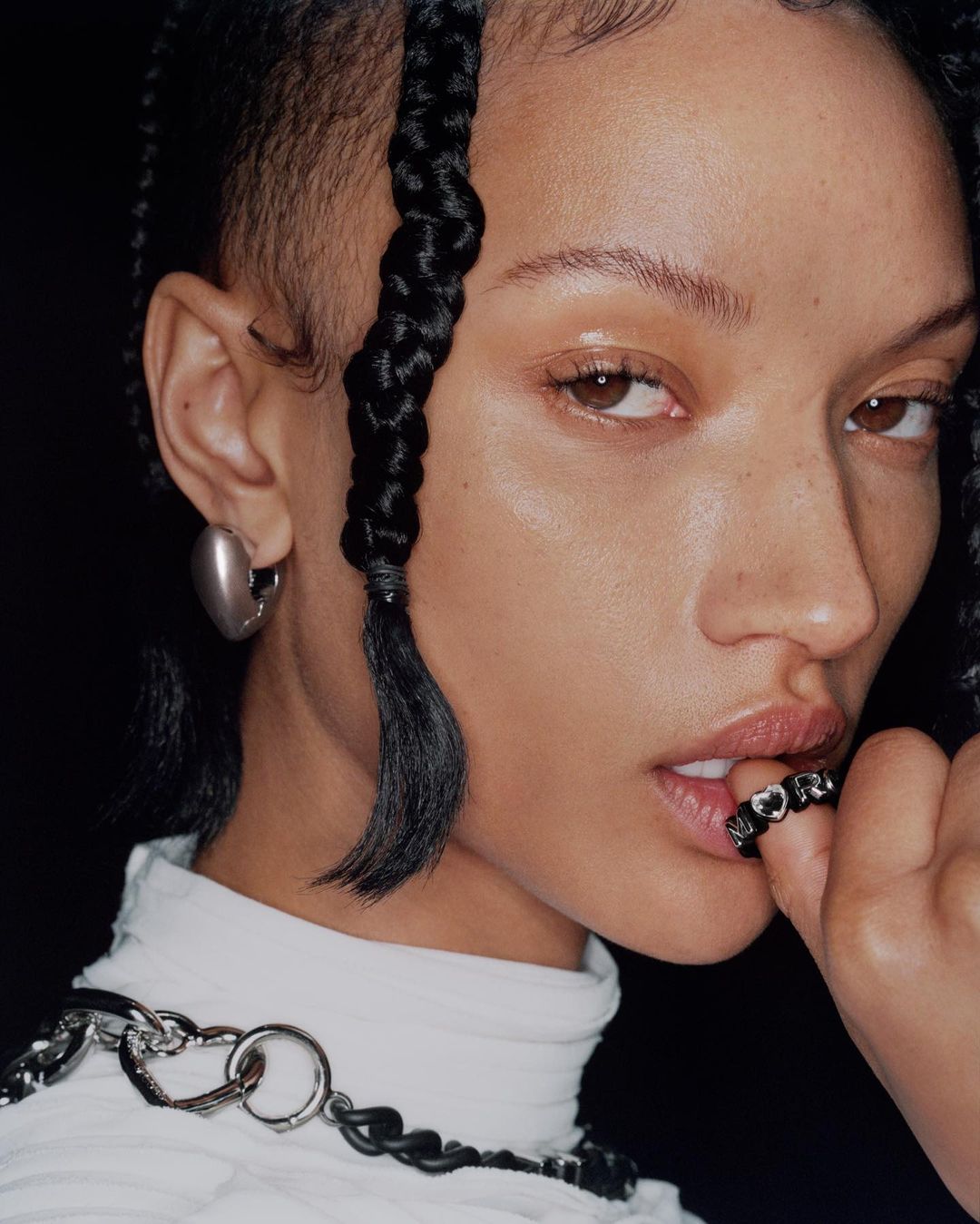 Close-up portrait of an adult woman with braided hair, wearing a white top, with a chain necklace and silver earrings against a black background.