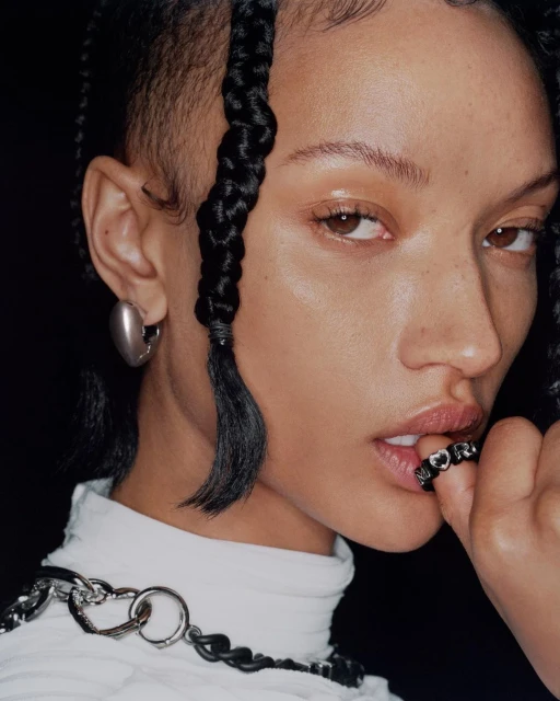 Close-up portrait of an adult woman with braided hair, wearing a white top, with a chain necklace and silver earrings against a black background.