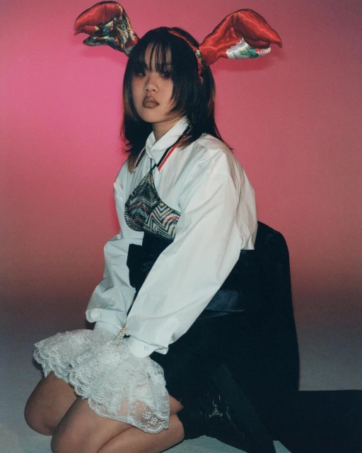 Young Asian woman with sculptural bunny ears, wearing a white shirt and a textured crop top, kneels against a mottled pink backdrop.