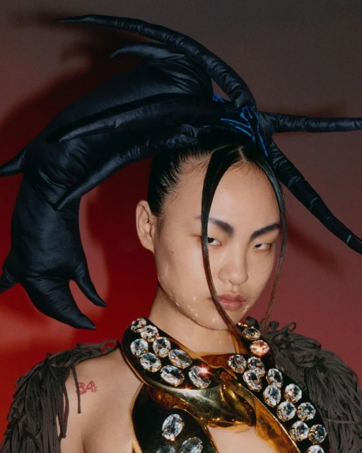 Asian woman in elaborate black headpiece and jewelry poses against a red background in a high-fashion editorial portrait.