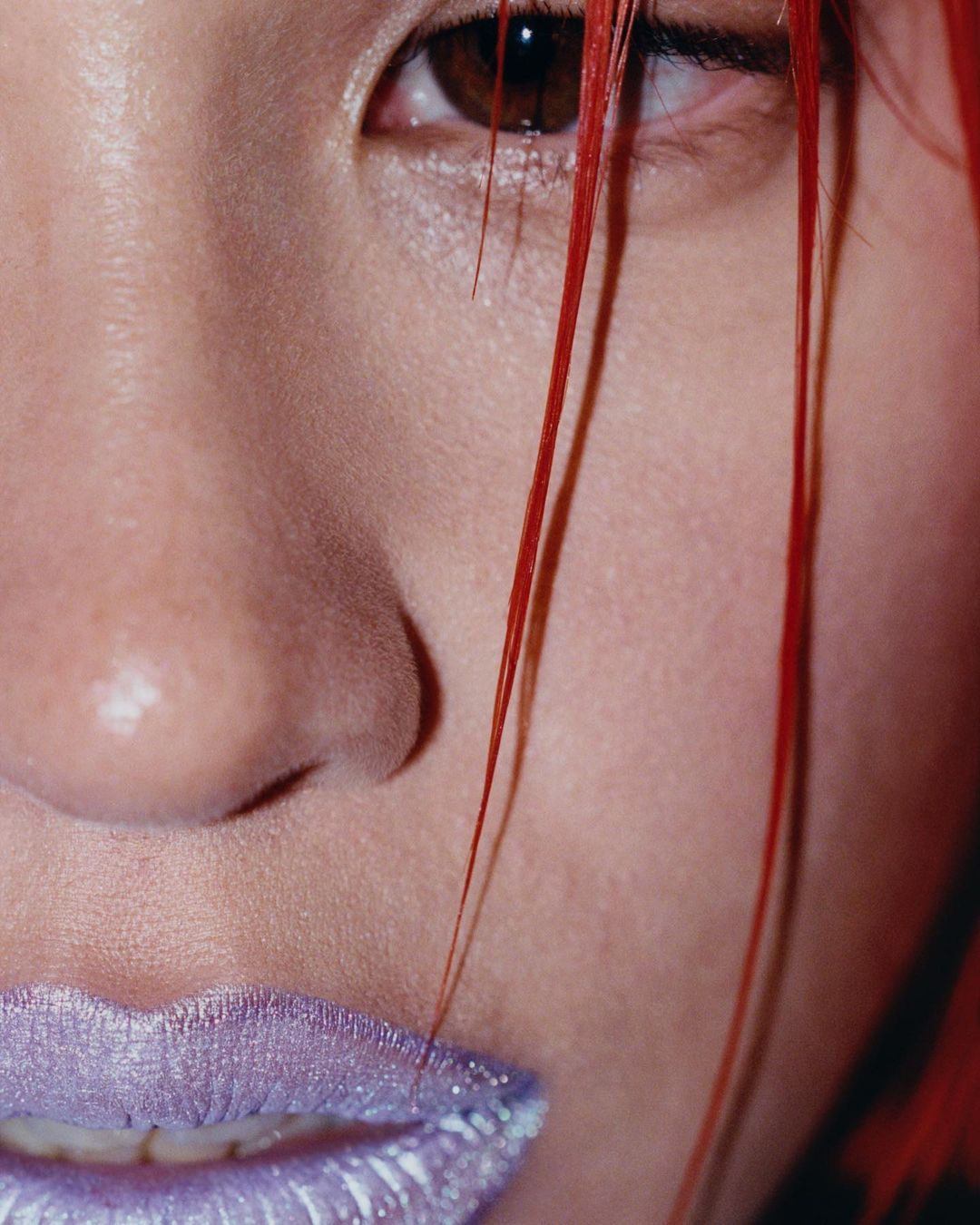 Close-up portrait of a woman's face with red hair partially covering one eye and wearing sparkling, iridescent purple lipstick.
