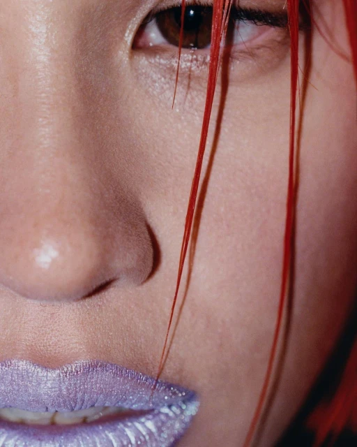 Close-up portrait of a woman's face with red hair partially covering one eye and wearing sparkling, iridescent purple lipstick.