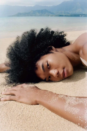 Close-up portrait of a man with dark curly hair lying on a sandy beach looking toward the camera.