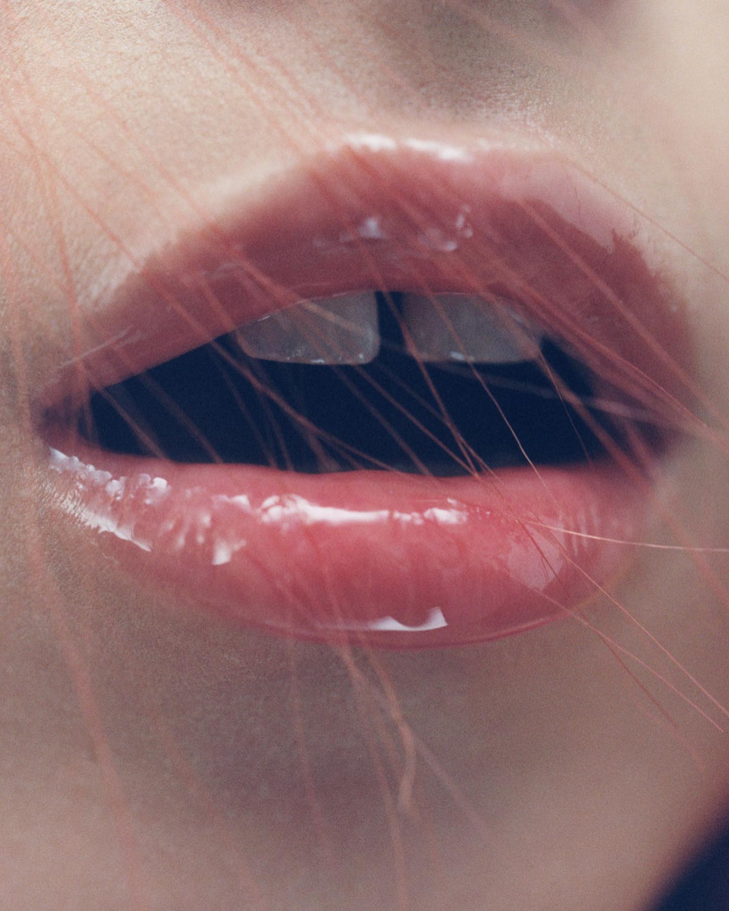 Close-up of a person's glossy lips with visible teeth and thin strands of hair or fiber crossing the foreground of the composition.