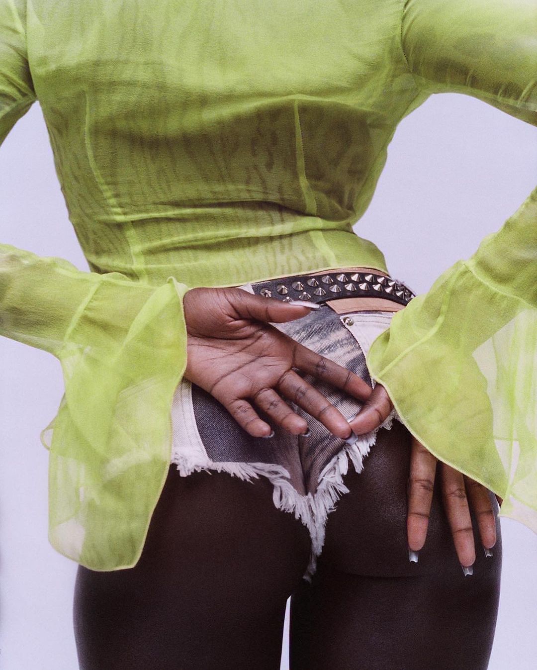 Back view of an African American adult female wearing a sheer chartreuse top, white frayed denim shorts, and a studded belt against a plain backdrop.