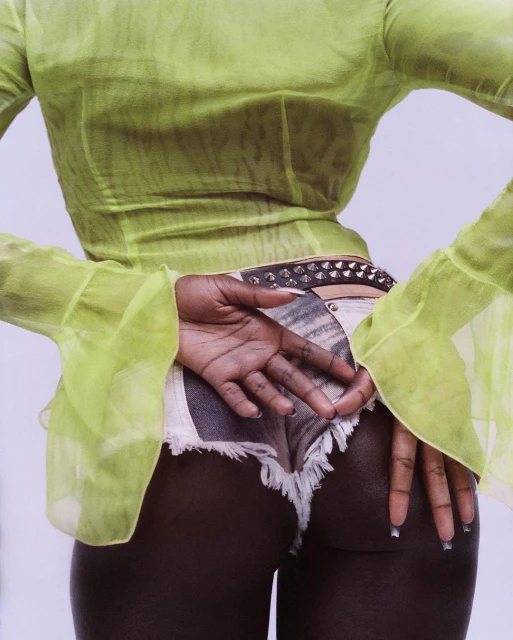 Back view of an African American adult female wearing a sheer chartreuse top, white frayed denim shorts, and a studded belt against a plain backdrop.