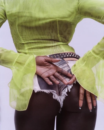 Back view of an African American adult female wearing a sheer chartreuse top, white frayed denim shorts, and a studded belt against a plain backdrop.