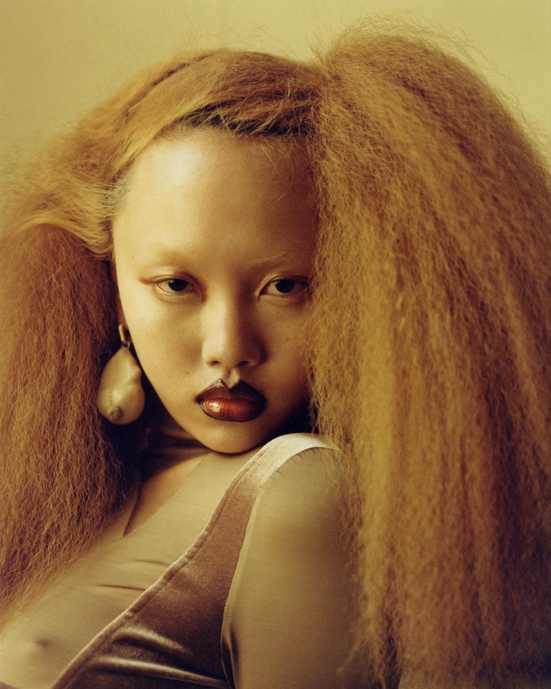 Close-up portrait of an Asian woman with distinctive makeup and voluminous orange hair wearing a taupe-colored top and statement earrings.