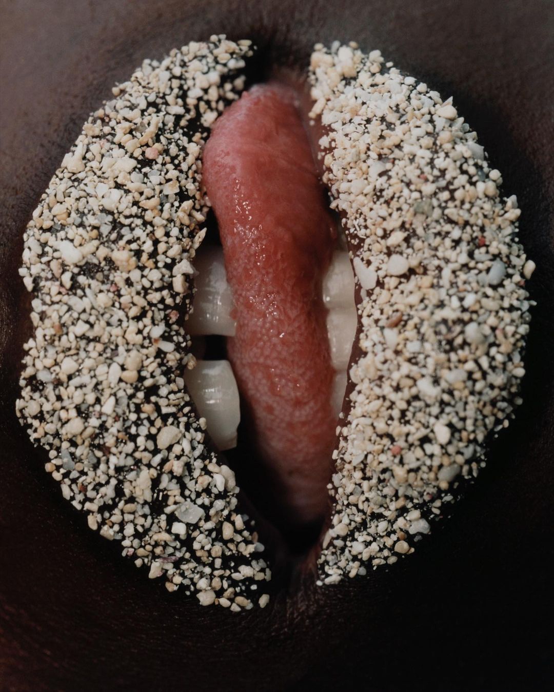 Close-up of open mouth with tongue and teeth visible, lips covered in granules, capturing an artistic and unusual beauty concept.