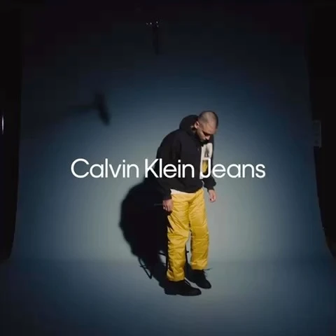 Man poses with Calvin Klein Jeans logo on a blue background, wearing a black hoodie, yellow pants, and black shoes in studio lighting.