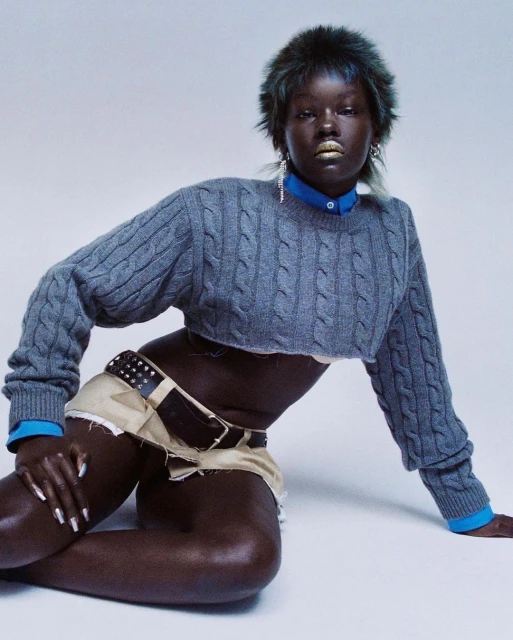 A dark-skinned woman with gold lipstick poses on a white background wearing a blue shirt, gray sweater, and a studded belt and khaki shorts.