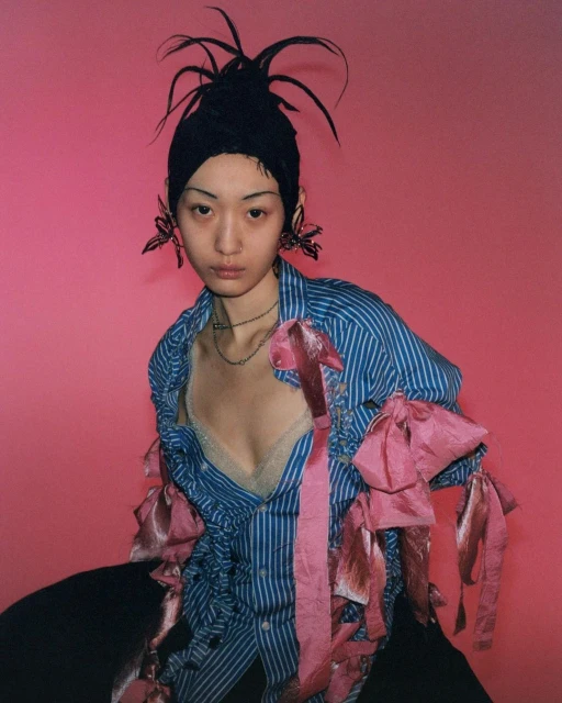 Woman with striking headpiece and striped blouse poses against a vibrant pink backdrop, showcasing a unique fashion aesthetic.