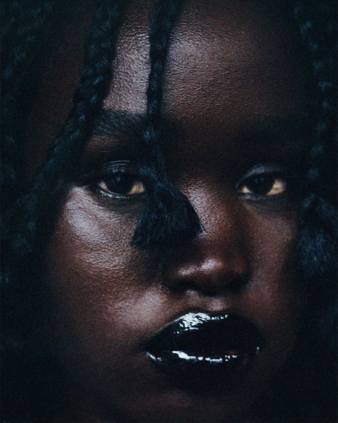 Close-up portrait of an adult female with dark skin, glossy black lips, and braided hair, set against a dark, indistinct background.